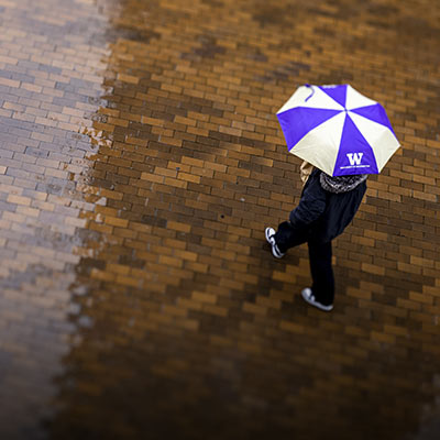 Student with a UW umbrella walks across Red Square in the rain.