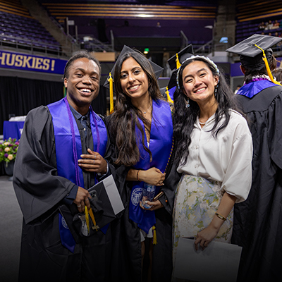 A trio of students, two of whom are dressed in graduation regalia, pose in an arena