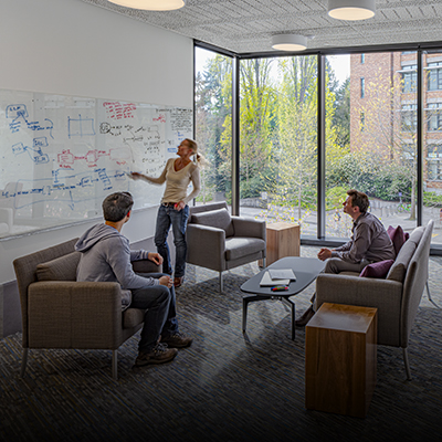 A group of three people, two of whom are seated listening to person standing and gesturing at a whiteboard