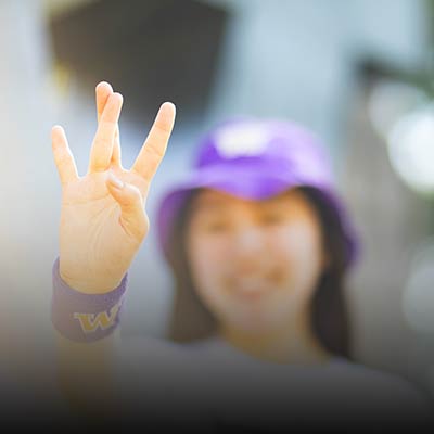 Female undergraduate student wearing a purple hat and giving the W (dubs up) hand sign.