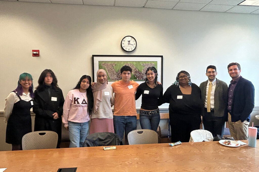 A group of seven college students stand, arms interlinked, alongside Armon Dadgar and Joshua Kalla in a conference room