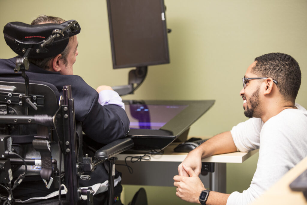 Accessible Technology Grand Challenge: A gentleman sitting in a wheelchair, uses a smart touchscreen while a Ph.D. student watches.