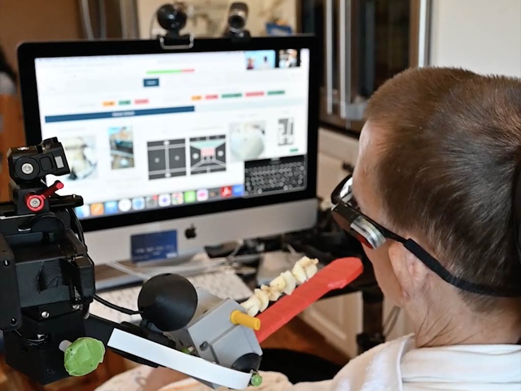 A man in bed and looking at a computer screen, tele-operating an assistive robot.