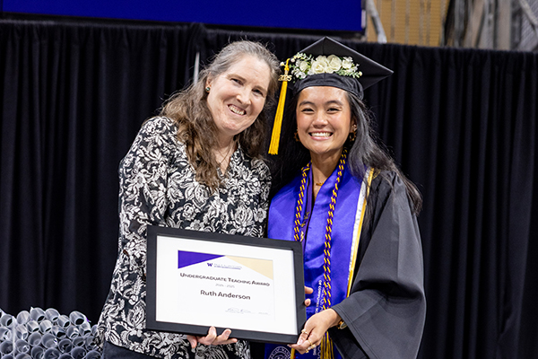 A woman with long hair wearing a patterned blouse stands next to a smiling younger woman dressed in graduation regalia jointly holding an award plaque in front of a table piled with rolls of commemorative diplomas