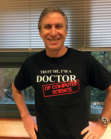 Hank Levy smiling showing off a t-shirt that says "Trust me I'm a Doctor of Computer Science"