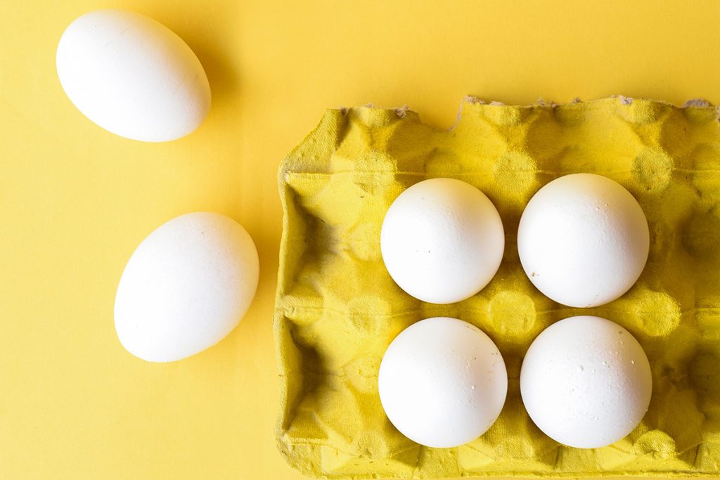 A yellow carton full of white eggs sits against a yellow background.