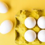 A yellow carton full of white eggs sits against a yellow background.