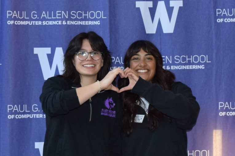 Two Allen School undergraduate students hold their hands together to make a heart shape in front of an Allen School photo backdrop