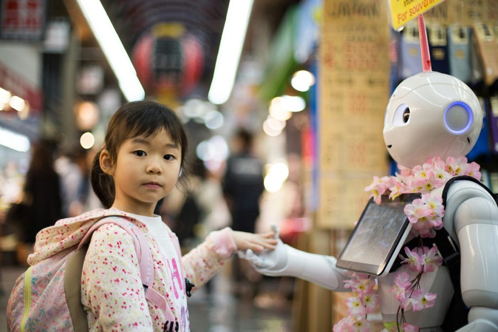 A small child wearing a hoodie and backpack holds hands with a robot humanoid.