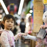 A small child wearing a hoodie and backpack holds hands with a robot humanoid.