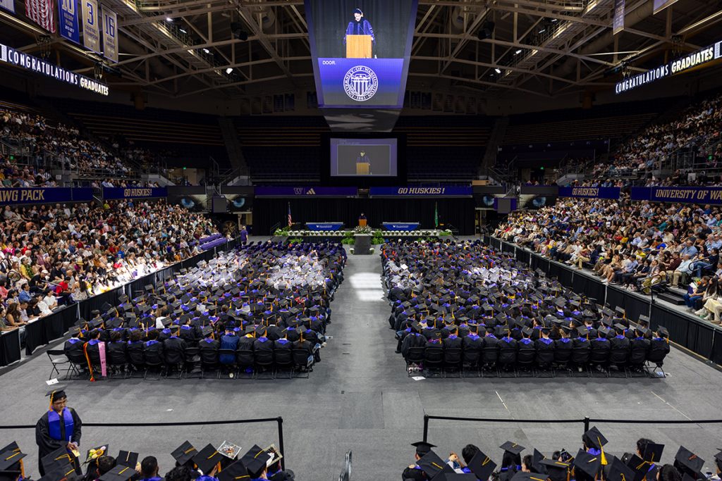 Wide angle photo of the arena with graduates in regalia seated on the floor surrounded by bleachers full of crowds beneath the jumbotron displaying a speaker in regalia