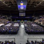 Wide angle photo of the arena with graduates in regalia seated on the floor surrounded by bleachers full of crowds beneath the jumbotron displaying a speaker in regalia
