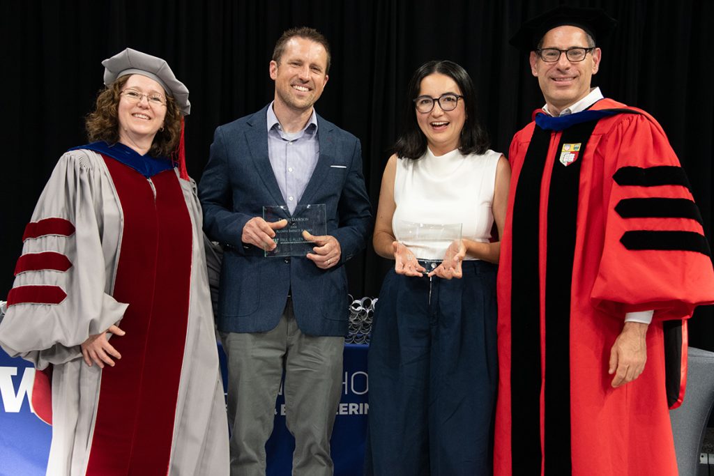 Two people dressed in business casual attire holding award plaques flanked by two people dressed in Ph.D. regalia from their respective alma maters