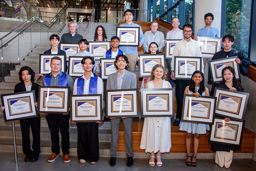 A group of smiling students and faculty pose on a staircase holding framed award certificates