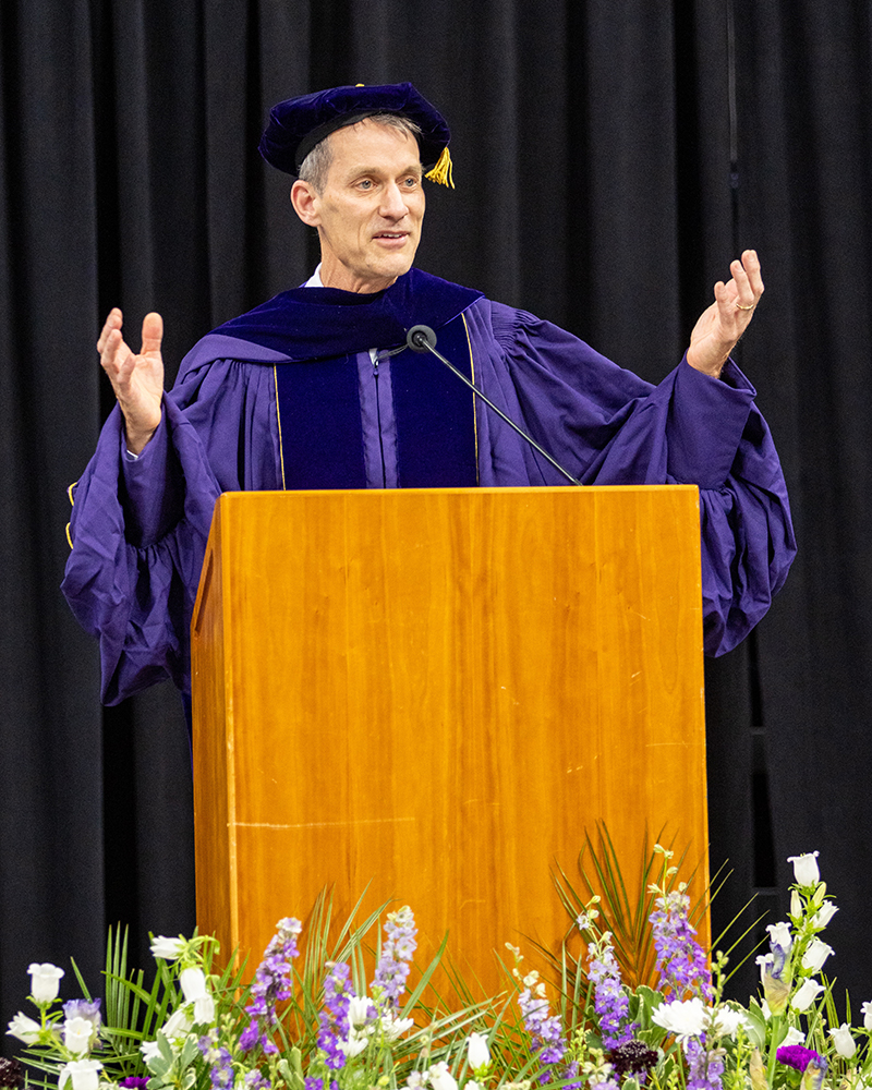 Jeff Dean in Ph.D. regalia raises his hands while addressing the graduates from the podium behind a row of flowers