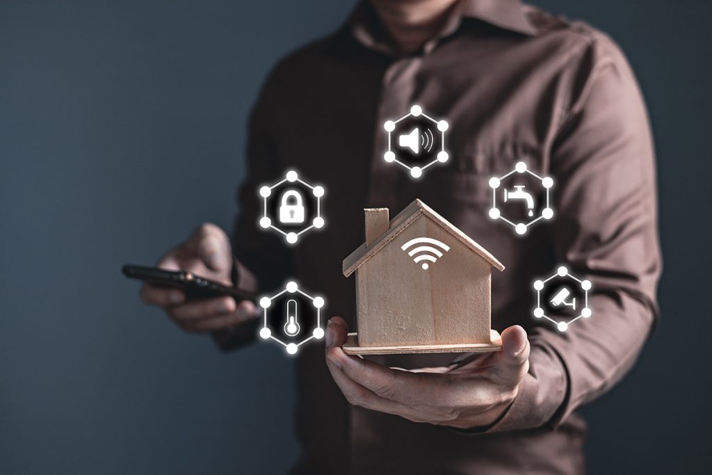 Man holding home model and software icons (temperature, sound, padlock, water, CCTV) controlling appliances smart home technology applications on smartphone screen.