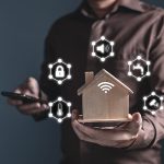 Man holding home model and software icons (temperature, sound, padlock, water, CCTV) controlling appliances smart home technology applications on smartphone screen.
