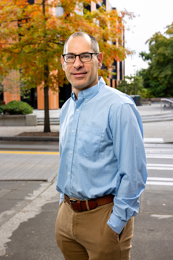 Dan Grossman in a relaxed stance with hands in pockets in front of the crosswalk approaching the Gates Center flanked by fall foliage