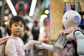 A small child wearing a hoodie and backpack holds hands with a robot humanoid.