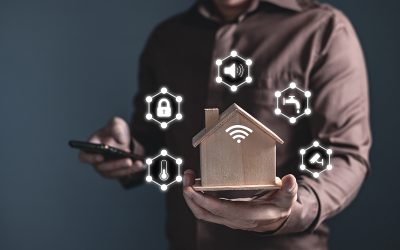 Man holding home model and software icons (temperature, sound, padlock, water, CCTV) controlling appliances smart home technology applications on smartphone screen.