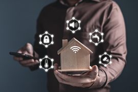 Man holding home model and software icons (temperature, sound, padlock, water, CCTV) controlling appliances smart home technology applications on smartphone screen.