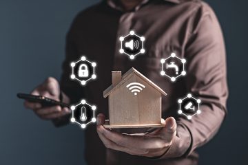 Man holding home model and software icons (temperature, sound, padlock, water, CCTV) controlling appliances smart home technology applications on smartphone screen.