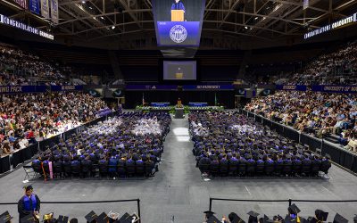 Wide angle photo of the arena with graduates in regalia seated on the floor surrounded by bleachers full of crowds beneath the jumbotron displaying a speaker in regalia
