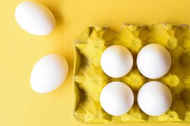 A yellow carton full of white eggs rests against a white background.