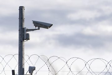 A surveillance camera mounted on a pole above a spiral of barbed wire fencing against a cloudy sky