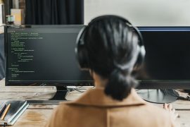 A person, viewed from behind, wearing headphones and looking at a pair of computer monitors