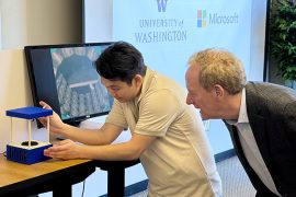 Brad Smith peers over a person's shoulder as they demonstrate a device that uses sound waves to levitate liquid droplets