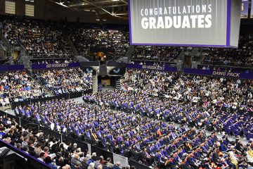 A college basketball arena decorated for graduation, with people wearing graduation regalia seated in rows of chairs on the carpeted floor, and people filling the stands to cheer them on. The jumbotron above the floor displays the message Congratulations, Graduates