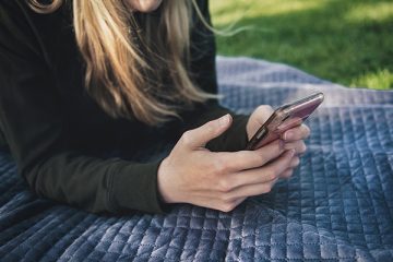 A young person resting on their elbows on a picnic blanket typing into a smartphone