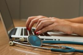 A person's hands typing on a laptop next to a stethoscope on a stable