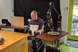 A person seated in a wheelchair at a table controls a laptop with a mouthpiece as a robotic arm spears a piece of food with a fork on a side table