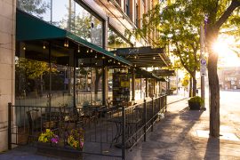 A sidewalk lined with trees alongside a cafe patio in the sunshine