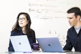 Su-In Lee sits at a table in front of a whiteboard with a graduate student behind a pair of open laptops