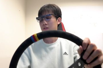 Xiyuan Alan Shen wearing goggles and seated behind the wheel of a driving simulator looks off to the side while participating in a study of distracted driving tied to touch screen use