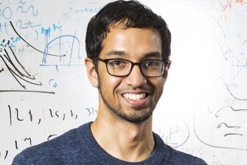 Portrait of Vikram Iyer smiling in front of a whiteboard displaying various handwritten diagrams, equations and notes